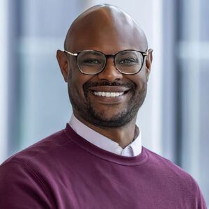 BMS employee Jamaal Sebastian-Barnes smiling, standing in an office setting, wearing eye glasses and a maroon colored sweater