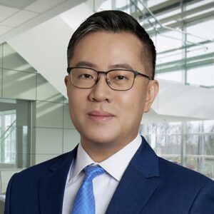 Headshot of BMS employee from China and Asia Cluster is in an office setting. He is smiling and wears glass with a dark suit and light blue tie.